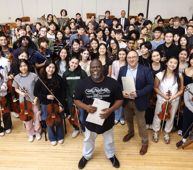 A group photo with Jeff Scott standing front and center with members of the Juilliard Pre-College Orchestra surrounding him. The group of young musicians is smiling and gathered in a rehearsal room with music stands and chairs in the background